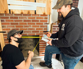 Two men are standing by a fireplace with a tape measure and a chimney inspection form.